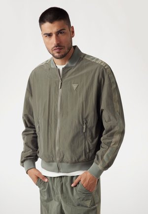 Lightweight olive green bomber jacket with zip closure, side pockets, and contrasting stripe detail featuring "GUESS" logo on sleeves.