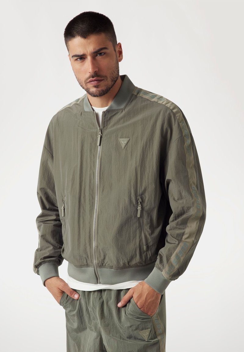 Cazadora bomber ligera en color verde oliva con cierre de cremallera, bolsillos laterales y detalle de franjas en contraste que presenta el logo "GUESS" en las mangas.
