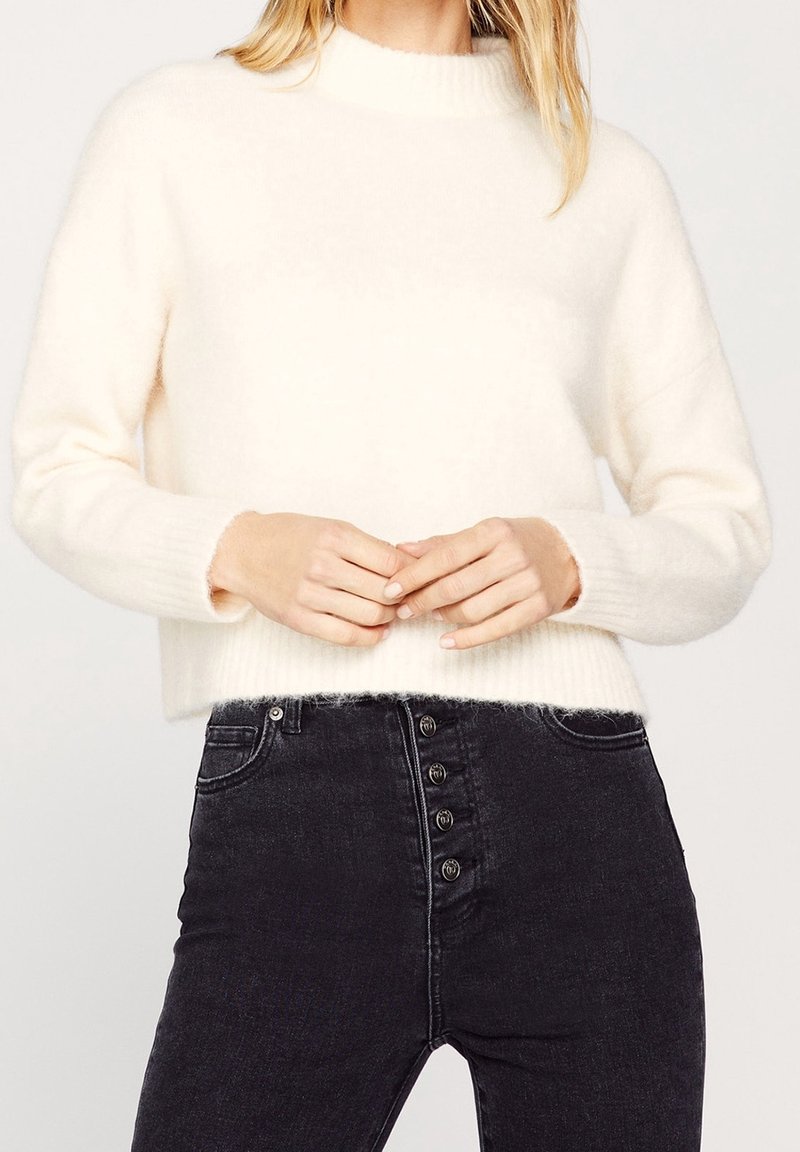 Femme portant un pull blanc duveteux et un jean noir taille haute avec des boutons sur le devant, mains jointes lâchement devant.