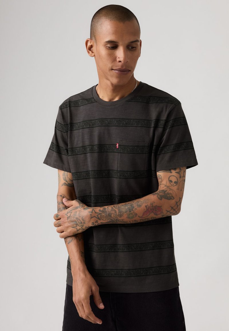 Homme tatoué portant un t-shirt noir rayé à manches courtes, regardant vers le bas, debout devant un fond clair uni.