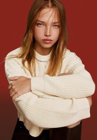 Krämfärgad kabelstickad tröja med rund halsringning och ribbade ärmslut. Modellens armar är korsade, vilket visar en mjuk textur mot en röd bakgrund.