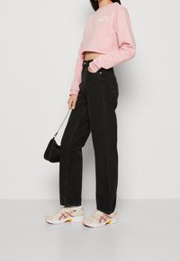 Felpa corta rosa con logo bianco, abbinata a jeans in denim nero a vita alta e sneakers beige arricchite da dettagli rossi e gialli.