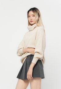 Beige gestrickter Rollkragensweater mit lockerer Passform, kombiniert mit einem dunkelgrauen, plissierten Minirock. Sanfte Textur und lässige Designdetails.