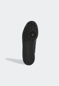 Black Adidas sneaker sole with circular tread patterns and Adidas logo in the center, casting a shadow on a white background.
