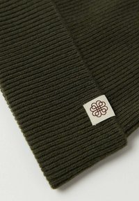 Mörkgrön stickad mössa med ribbat mönster och en liten tygetikett med en dekorativ logotyp i brunt.