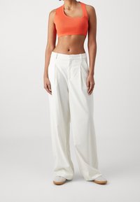 Kobieta ubrana w dopasowany pomarańczowy crop top, luźne białe spodnie z wysokim stanem i białe tenisówki na gumowej podeszwie, stojąca na jednolitym tle.