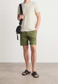 T-shirt beige a maniche corte con logo, pantaloni corti verde oliva, borsa a tracolla nera e ciabatte nere. Tessuto liscio senza motivi visibili.