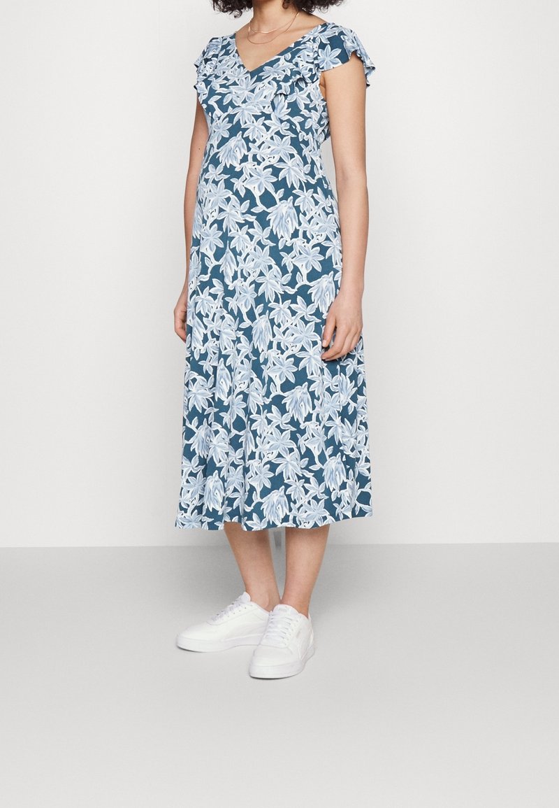 Femme portant une robe midi bleue à imprimé floral blanc et des baskets blanches, debout sur un fond clair uni.