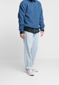 Blaues Fleece-Sweatshirt aus strukturiertem Stoff, über ein dunkles kariertes Hemd getragen, kombiniert mit hellblauen Jeans und weißen Sneakern.