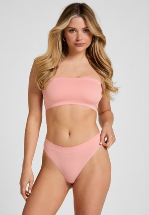 Vrouw met lang blond haar die een lichtroze strapless bandeautopje en bijpassende hoog uitgesneden broekjes draagt tegen een effen achtergrond.