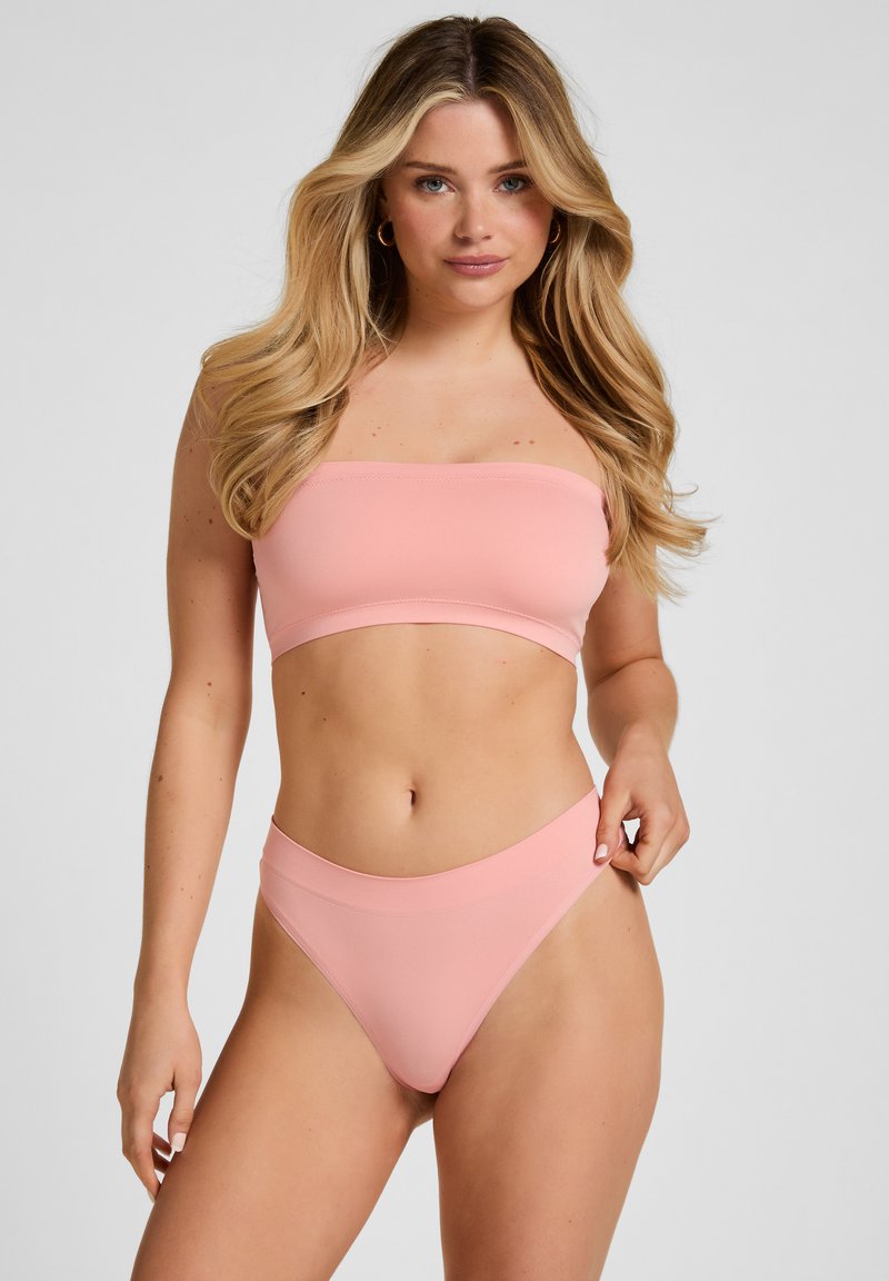 Donna con lunghi capelli biondi che indossa un top a fascia rosa chiaro senza spalline e slip abbinate a vita alta su uno sfondo semplice.