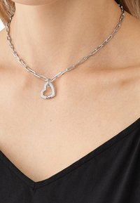 Collier en chaîne argentée avec de gros maillons et un pendentif en forme de cœur incrusté de diamants, porté sur un haut noir à col en V.