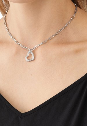 Collier en chaîne argentée avec de gros maillons et un pendentif en forme de cœur incrusté de diamants, porté sur un haut noir à col en V.