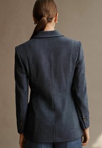 Žena s hnědými vlasy v culíku, oblečená v přizpůsobeném tmavě šedém sáčku, stojí zády k fotoaparátu na jednobarevném pozadí.