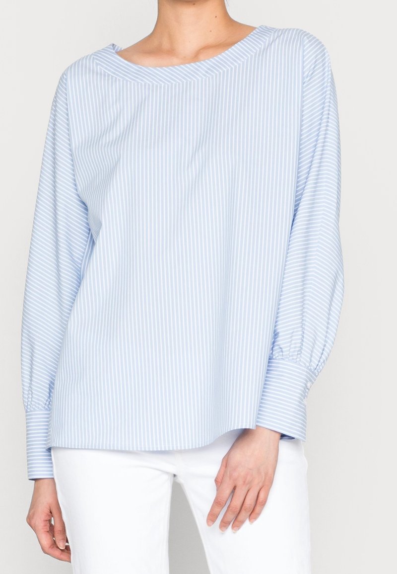Blouse à manches longues rayée verticalement bleu clair et blanc avec de larges poignets, portée avec un pantalon blanc sur une personne debout.
