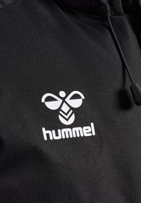 Schwarzer Hoodie mit strukturierter Oberfläche und einem markanten weißen Hummel-Logo mit Bienendesign, dazu Kordelzüge und ein schlanker Schnitt.