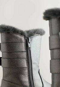 Close-up van metallic winterlaarzen met een zijrits en grijze nepbontvoering rond de bovenranden tegen een effen achtergrond.