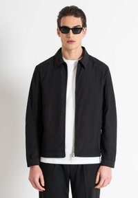 Giacca leggera nera con zip frontale, colletto a punta e maniche lunghe; abbinata a una maglietta bianca e pantaloni neri.