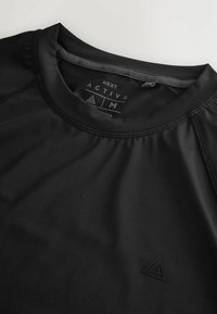 Czarna koszulka sportowa z okrągłym dekoltem, gładką fakturą i płaskimi szwami. Posiada małe czarne logo na klatce piersiowej.