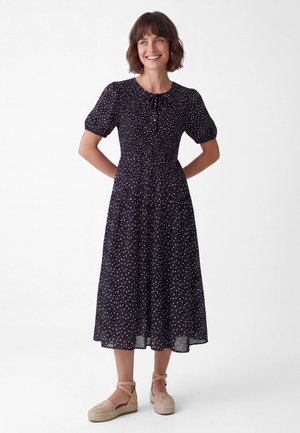 Smilende kvinde med kort brunt hår iført sort midi-kjole med hvide prikker og beige espadrilles, står med hænderne på ryggen.