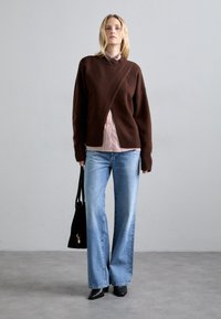 Rjav pleten pulover s prečnim dizajnom, nošen čez rdečo in belo prugasto bluzo. V kombinaciji z svetlo modrimi širokimi hlačami in črnimi čevlji.