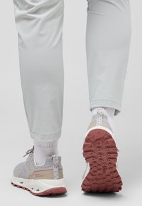 Des chaussures de randonnée gris clair présentent un dessus texturé, des matériaux synthétiques, une semelle blanche et une semelle en caoutchouc rouge. Les accents comprennent une languette à motif.