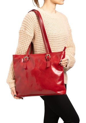 Rode leren tote bag met glanzende afwerking, twee bijpassende hengsels en metalen gespen. Heeft een ruime indeling en wordt vastgehouden door een persoon in een trui.