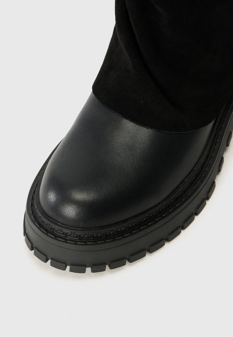 Schwarzer Knöchelstiefel mit einer glatten Lederzehe, einem Obermaterial aus Wildleder und einer dicken Profilsohle mit dekorativen Nähten am Rand.
