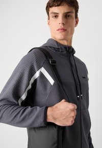 Pilkas raštuotas zip-up džemperis su tekstūruotu audiniu, baltomis įstrižomis juostelėmis, ir juoda rankinė su petnešomis, kurią laiko asmuo.