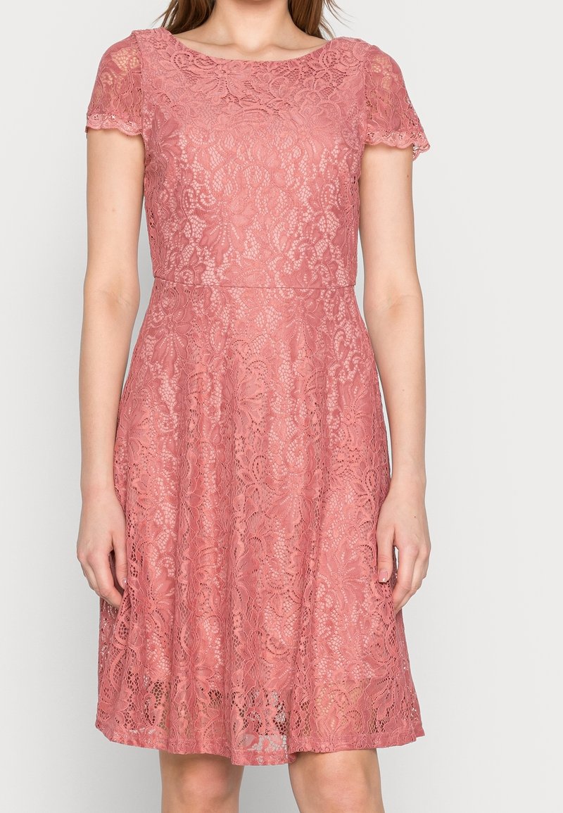 Femme portant une robe en dentelle rose poudré à manches courtes et taille ajustée, debout devant un fond clair uni.