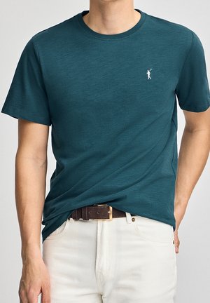 Grønblå bomuld t-shirt med rund hals og korte ærmer, der har en hvid logo-accent på brystet. Båret med hvide bukser og et brunt bælte.