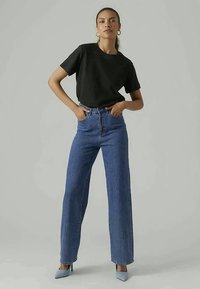 Woman wearing black T-shirt, high-waisted blue jeans, and light blue heels standing against plain gray background with hands in pockets.