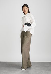 White blouse with voluminous sleeves, paired with high-waisted brown plaid trousers and sleek white heels; holding a small black clutch.