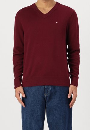 Krepisierter burgunderfarbener V-Ausschnitt-Pullover aus Strickmaterial, mit langen Ärmeln und geripptem Saum. Kleines Logo auf der linken Brust. Getragen mit blauen Jeans.