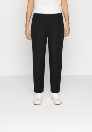 Personne portant un pantalon droit noir avec un bouton et une taille élastique, associé à des baskets blanches et un haut blanc sans manches.