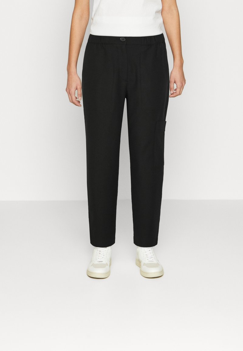 Personne portant un pantalon droit noir avec un bouton et une taille élastique, associé à des baskets blanches et un haut blanc sans manches.