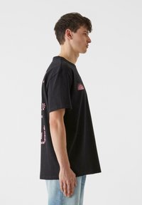 Must must tshirt lühikeste varrukatega, roosa graafikaprindiga, ümmarguse kaelusega ja lõdva lõikega. Kujutatud külje pealt.