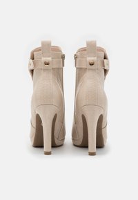 Bottines beige à talons hauts avec motif texturé, fermeture éclair arrière et brides décoratives avec boutons dorés, vues de dos.