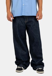 Dark blue denim jeans with a loose fit, featuring visible stitching and a straight leg design. Paired with black sneakers.
