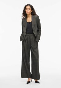 Sivi karirani vuneni kostim koji sadrži moderno krojen sako i hlače širokih nogavica. Glatka tekstura s suptilnim narančastim akcentima; nošen s crnom majicom i potpeticama.