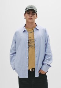 Blå og hvid stribet skjorte med lange ærmer over en beige grafisk t-shirt. Sorte jeans og en grå kasket fuldender outfittet.