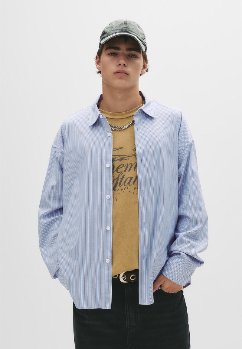 Blå og hvid stribet skjorte med lange ærmer over en beige grafisk t-shirt. Sorte jeans og en grå kasket fuldender outfittet.