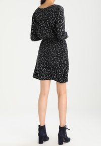 Czarna sukienka w wzory, z długimi rękawami, dopasowaną talią i rozkloszowanym dołem. Do tego granatowe botki na obcasie do kostki. Tło jest neutralne.