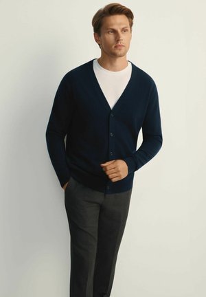 Granatowy kardigan wykonany z miękkiej dzianiny, z dekoltem w serek, pięcioma guzikami z przodu i ściągaczami przy mankietach, w połączeniu z szarymi spodniami.