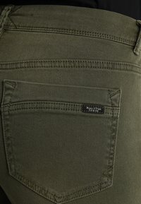 Jeans de mezclilla verde oliva con un bolsillo trasero que presenta una etiqueta de marca negra. La tela tiene una textura suave y resistente, con detalles de costura prominentes.