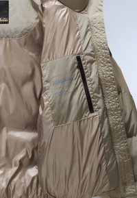 Interno della giacca puffer beige che mostra una tasca con zip con il logo del marchio stampato e un'etichetta che afferma "Questo puffer è 100% senza piuma."