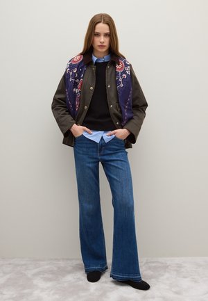 Giacca marrone con sciarpa ricamata blu scuro, maglione nero, camicia azzurro chiaro e jeans blu a gamba larga. Tessuto testurizzato, accenti floreali.