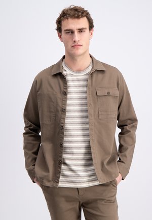 Ung mand iført brun knappet jakke over en beige og gråstribet skjorte med hænderne i lommerne, foran en ensartet lys baggrund.