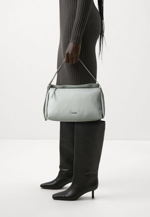 Personne en robe côtelée tenant un sac à main gris clair, portant des bottes en cuir noires à talons hauts, se tenant devant un fond clair uni.