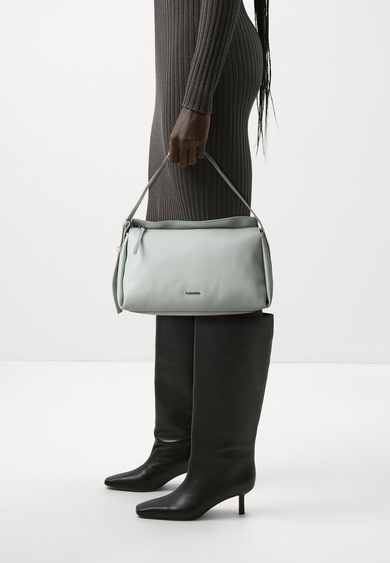 Personne en robe côtelée tenant un sac à main gris clair, portant des bottes en cuir noires à talons hauts, se tenant devant un fond clair uni.